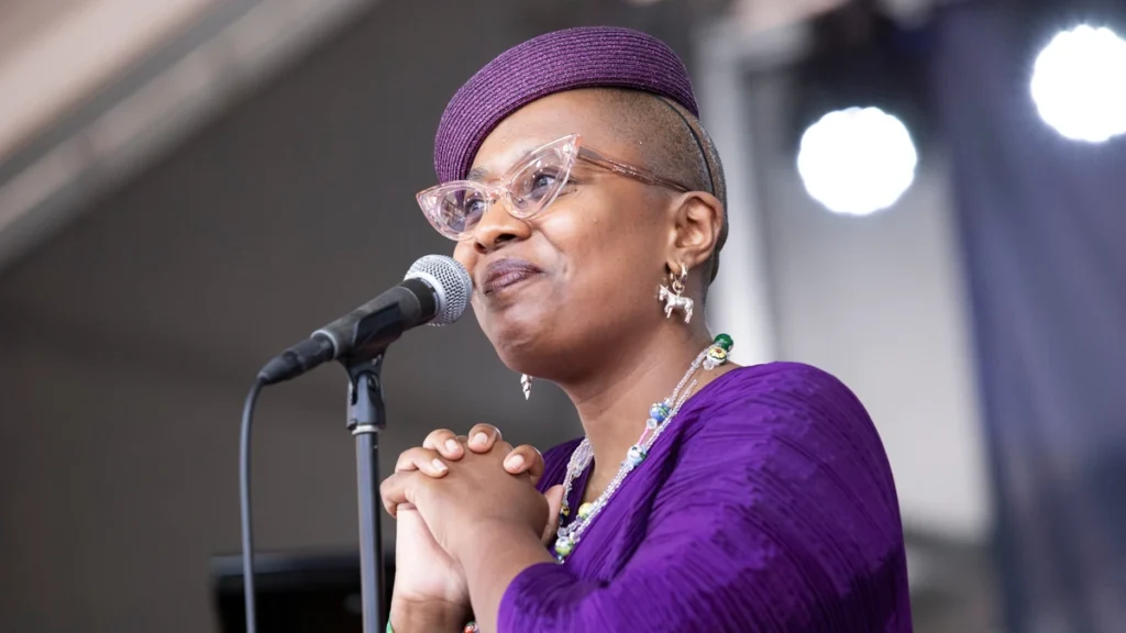 Cecile McLorin Salvant singing at a microphone in a purple dress and hat, hands clasped