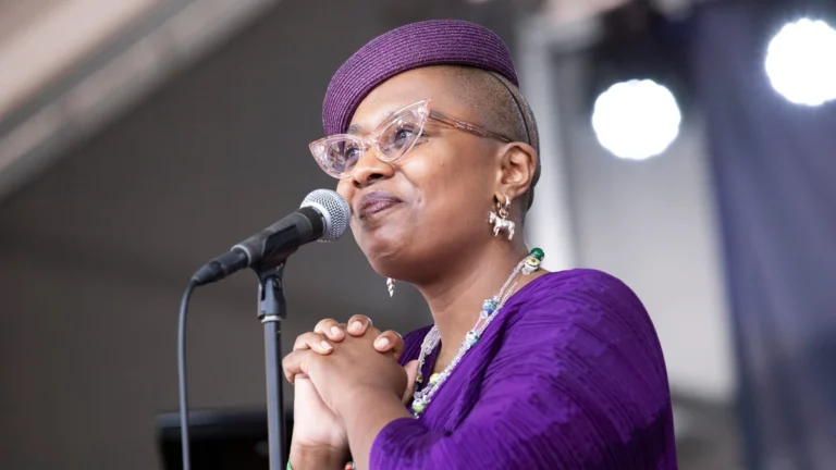 Cecile McLorin Salvant singing at a microphone in a purple dress and hat, hands clasped